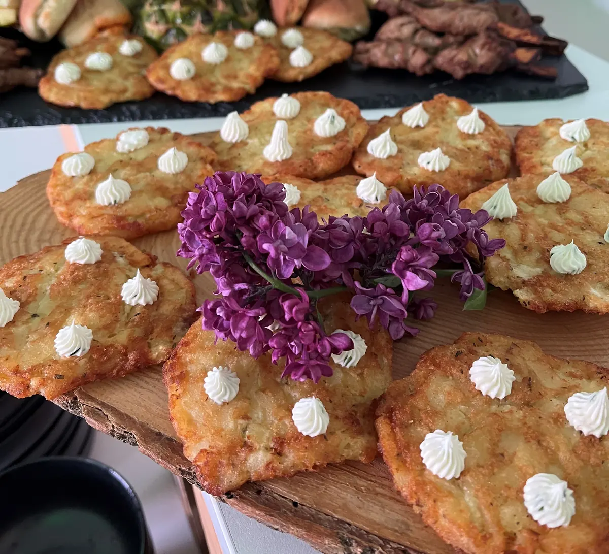 Kartoffelpuffer mit Cremetupfen und Fliederblüten auf einem Holzbrett, dahinter ein Buffet